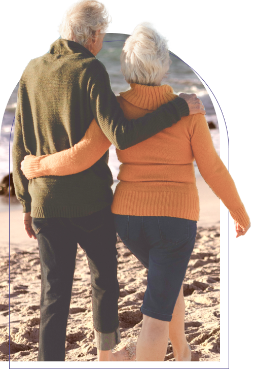 Elderly couple walking on the beach with their arms behind each others backs.